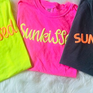 May include: Five different t-shirts with summer-themed text in various colors. The shirts are laid out on a white surface. The text on the shirts reads: "Summer Lovin", "Sunkissed", "Sunkissed", "Sunkissed", and "Salty".