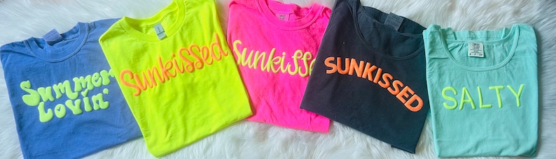 May include: Five different t-shirts with summer-themed text in various colors. The shirts are laid out on a white surface. The text on the shirts reads: "Summer Lovin", "Sunkissed", "Sunkissed", "Sunkissed", and "Salty".