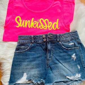 May include: A pink tank top with the word "Sunkissed" in yellow embroidery. The tank top is paired with blue denim shorts that have distressed details.