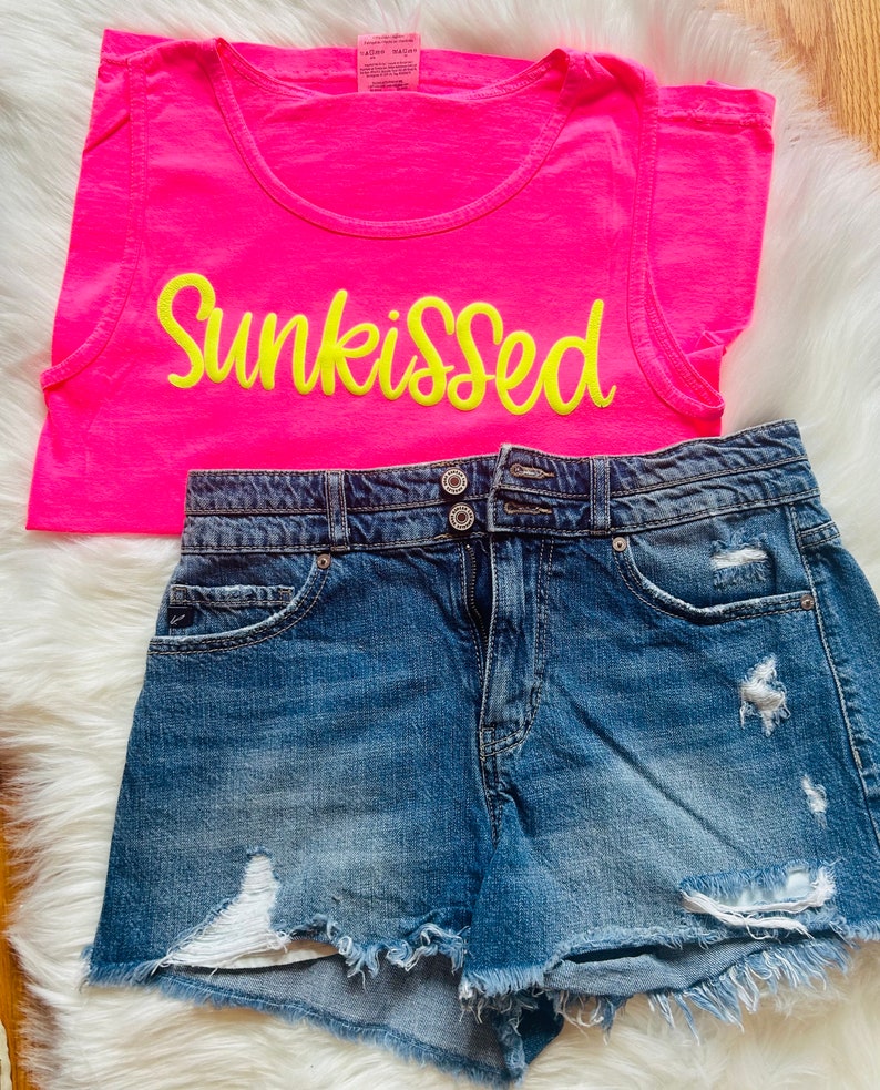 May include: A pink tank top with the word "Sunkissed" in yellow embroidery. The tank top is paired with blue denim shorts that have distressed details.
