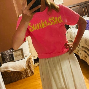 May include: A pink t-shirt with the word "Sunkissed" written in yellow script. The person wearing the shirt is also wearing a white skirt.
