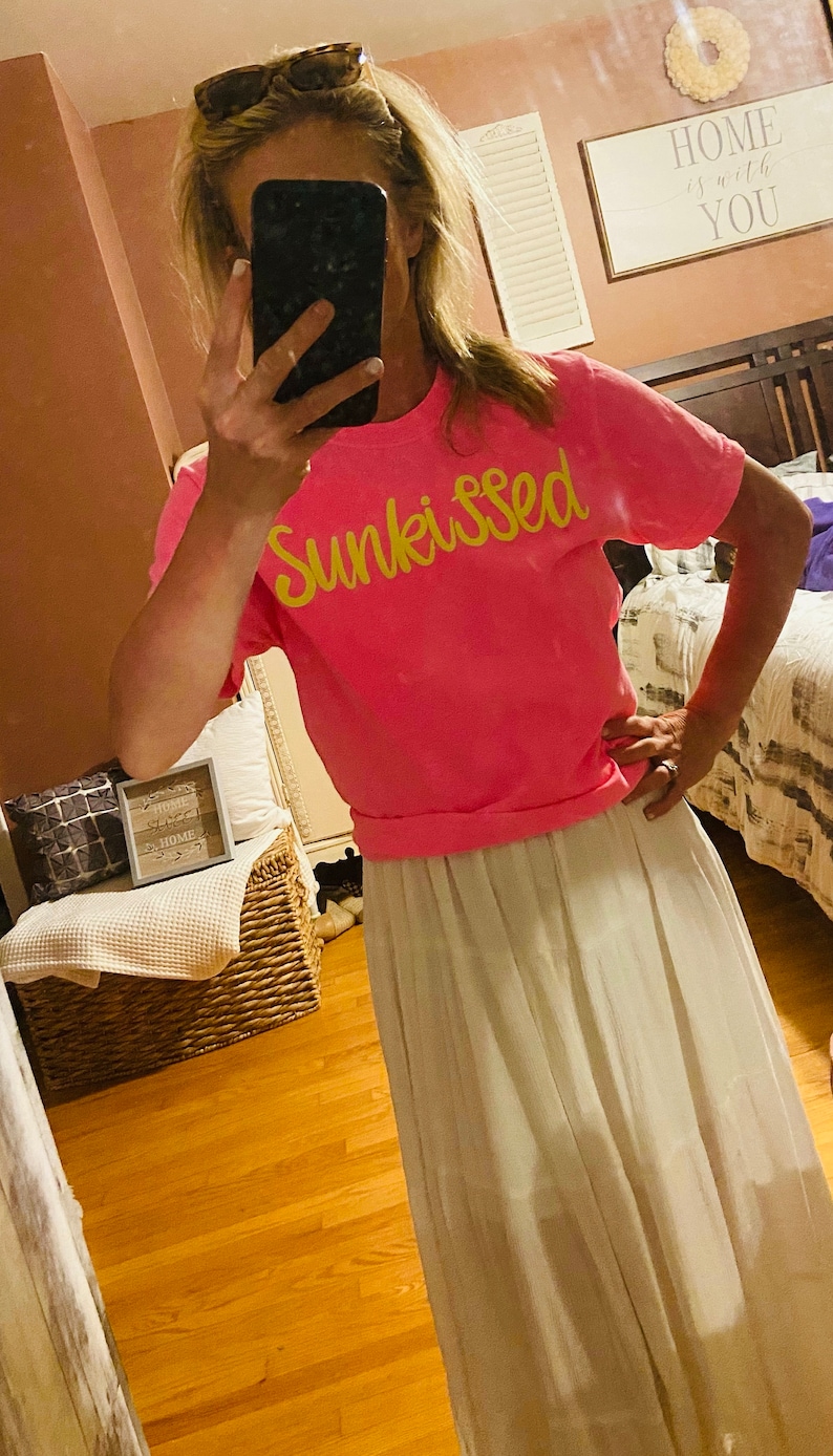 May include: A pink t-shirt with the word "Sunkissed" written in yellow script. The person wearing the shirt is also wearing a white skirt.