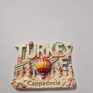 May include: A souvenir magnet featuring the word "TURKEY" in beige, with a landscape of Cappadocia. The magnet includes a red and yellow hot air balloon and the word "Cappadocia".