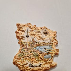 May include: A detailed souvenir magnet in the shape of Kuwait. The magnet depicts the city skyline, a compass rose, and the word "Kuwait". The colour palette includes shades of brown, beige, green, and blue. The magnet is a travel-themed item.
