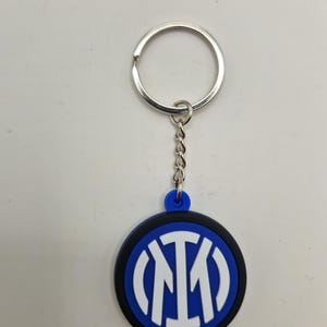 May include: A round keychain featuring the Inter Milan logo. The logo is a white "IM" design inside a blue circle, with a black border. The keychain has a silver key ring and chain.