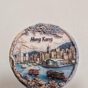 May include: A round, 3D, ceramic souvenir magnet depicting the Hong Kong skyline in detail. The magnet is painted in shades of blue, brown, and white, with the text "Hong Kong" printed in black at the top.