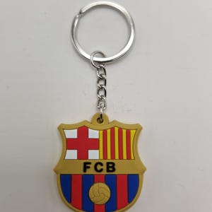 May include: A keychain featuring the FC Barcelona crest. The crest is shield-shaped with a gold border, and includes a red cross, yellow and red stripes, and a soccer ball. The letters "FCB" are in black.