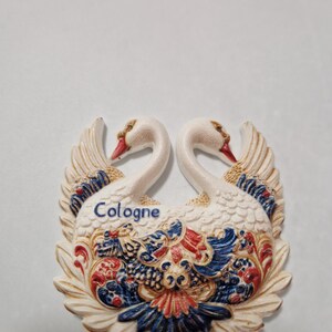 Puede incluir: Un imán decorativo con dos cisnes enfrentados. Los cisnes tienen cuerpos blancos, picos rojos y alas detalladas con detalles azules, rojos y dorados. La palabra "Cologne" está escrita en azul.