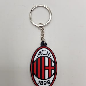 May include: Keychain with the ACM Milan logo. The oval charm has a black border, red and black stripes, a red cross, and the text "ACM" and "1899" in white. It features a silver metal ring and chain, suitable for keys or bags.