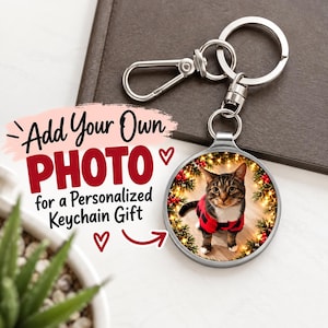 Op de afbeelding: Een ronde zilverkleurige metalen sleutelhanger met een aangepaste ronde foto-inzet van een bruine cyperse kat in een rood-zwart geruit hemd, omringd door een feestelijke krans van dennentakken en rode bessen met warme lichtjes.