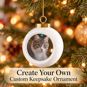 Op de afbeelding: Een rond wit keramieken ornament met een ronde uitsparing waarin een foto van een bruin-witte cyperse kat is geplaatst. Het ornament heeft een goudkleurige metalen dop en een gouden ophangkoord. De tekst luidt: Create Your Own Custom Keepsake Ornament.