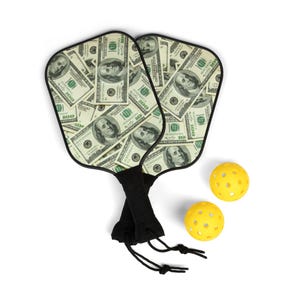 May include: Two black pickleball paddles with a design of US dollar bills. The paddles are on a white background with two yellow pickleball balls.