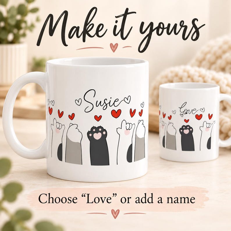 Op de afbeelding: Twee witte keramische mokken met een afbeelding van opgeheven kattenpootjes en rode hartjes. De grotere mok toont de naam Susie, terwijl de kleinere mok het woord love bevat. De tekst op de afbeelding luidt Make it yours en Choose Love or add a name.