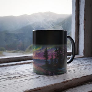 May include: Black coffee mug with a landscape design featuring a colorful sky, trees, and water. The mug is sitting on a wooden windowsill with a mountain view in the background. Steam rises from the mug.