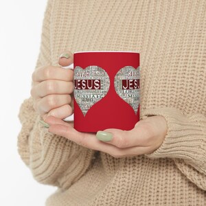 May include: A white ceramic mug with a red heart design. The heart shapes contain the word "JESUS" and other religious words. The mug is held by a person.
