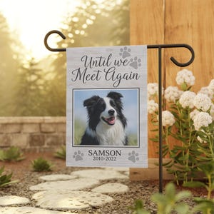 Op de afbeelding: Een gedenkteken tuin vlag met een zwart-wit hondenportret en de tekst "Until we Meet Again". De vlag bevat de naam van de hond, "Samson", en de data, 2010-2022, met pootafdruk accenten. De vlag wordt getoond op een zwarte metalen standaard.