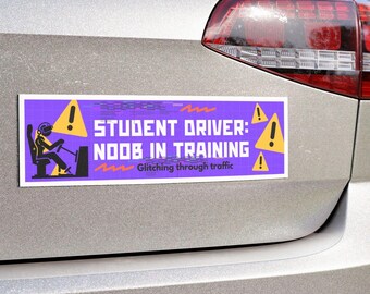 Pode incluir: Um autocolante retangular para pára-choques com fundo roxo e o texto "STUDENT DRIVER: NOOB IN TRAINING - Glitching through traffic." O autocolante também apresenta um boneco a conduzir um carro e símbolos de ponto de exclamação amarelos.