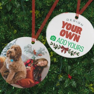 Può includere: Due decorazioni natalizie rotonde e bianche con nastri rossi. Una presenta una foto di un cane e una persona. L'altra ha il testo "DESIGN YOUR OWN ADD YOURS" in rosso e verde, con agrifoglio festivo.