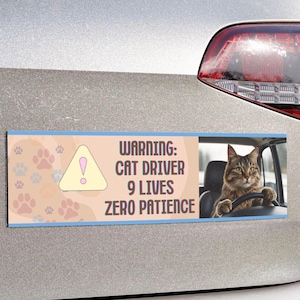 May include: A rectangular car bumper sticker with a light blue border and a peach background. The left side features paw prints and a yellow warning sign. The right side shows a cat driving a car. Text reads: "WARNING: CAT DRIVER 9 LIVES ZERO PATIENCE."