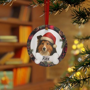 Può includere: Ornamento natalizio rotondo con un cane con un cappello da Babbo Natale. Il ritratto del cane è centrato su uno sfondo bianco, con un elemento a forma di osso sottostante con il nome "Rex". L'ornamento ha un bordo a scacchi rosso e nero con pigne e vegetazione.