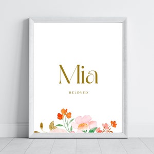 Peut inclure: Une impression encadrée blanche avec le nom "Mia" en lettres dorées et le mot "BELOVED" en dessous. Le bas de l'impression présente une illustration aquarelle de fleurs roses et orange.