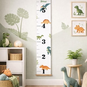 May include: A vertical white canvas growth chart hanging from a wooden frame, featuring black numerical markings from 1 to 5 and colorful illustrations of various dinosaurs, with the word Name printed in green at the top.