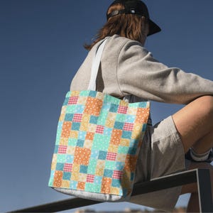 May include: A colorful tote bag with a patchwork design. The bag features squares of orange, blue, yellow, and pink with heart, dot, and gingham patterns. The bag has white straps and is carried over the shoulder.