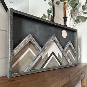 May include: A framed wood wall art piece featuring a geometric mountain design with a white circle moon. The mountains are made of wood with a distressed gray, white, and brown paint finish. The frame is black.