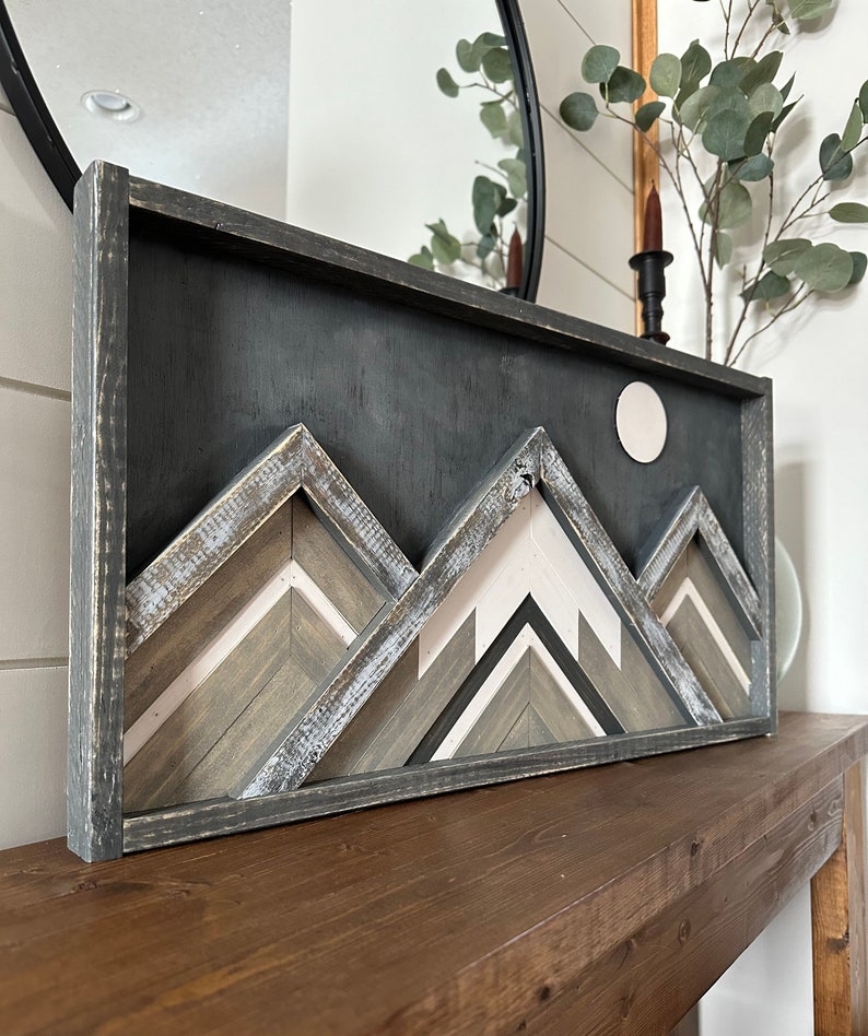 May include: A framed wood wall art piece featuring a geometric mountain design with a white circle moon. The mountains are made of wood with a distressed gray, white, and brown paint finish. The frame is black.
