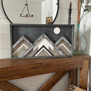 May include: A wooden mountain range wall art with a white moon and a distressed gray finish. The art is framed in a dark gray frame and is displayed on a wooden console table.