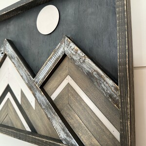 May include: A framed wood wall art piece with a black background and a white circle in the upper center. The piece features a geometric design of three mountains in shades of gray and white.