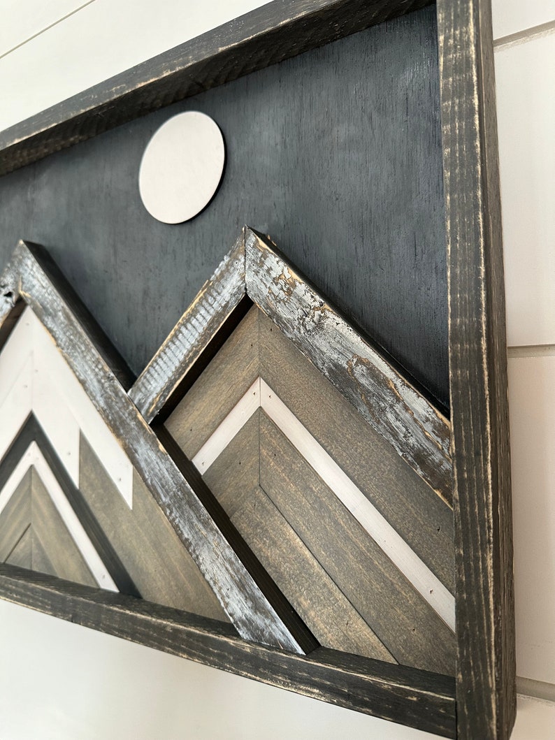 May include: A framed wood wall art piece with a black background and a white circle in the upper center. The piece features a geometric design of three mountains in shades of gray and white.