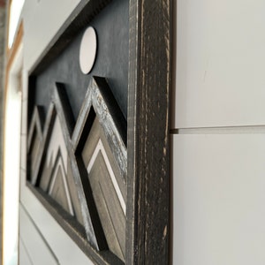 May include: A black framed wall art piece with a geometric mountain design in shades of gray and white. The mountains are layered and have a 3D effect. A white circle is in the upper center of the piece.