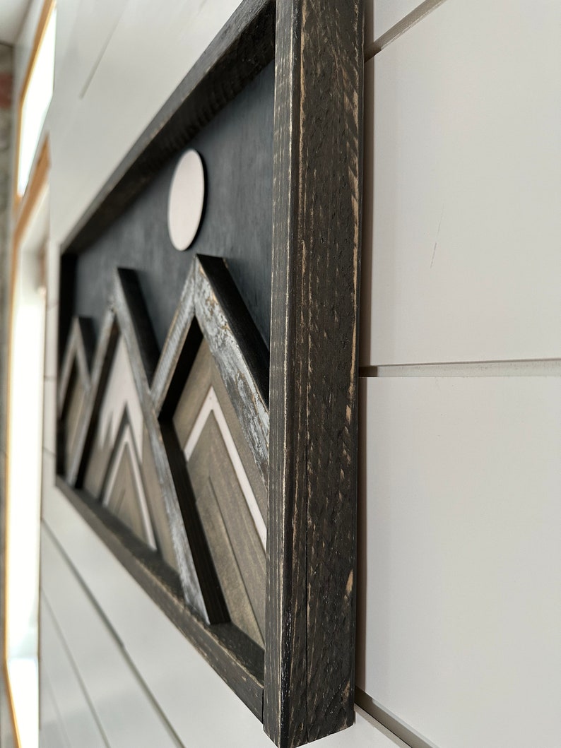 May include: A black framed wall art piece with a geometric mountain design in shades of gray and white. The mountains are layered and have a 3D effect. A white circle is in the upper center of the piece.