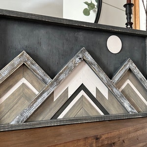 May include: A framed wooden wall art piece featuring a geometric mountain range design in shades of gray, white, and brown. The mountains are layered and textured, creating a three-dimensional effect. A white circle is centered at the top of the piece, representing the moon.
