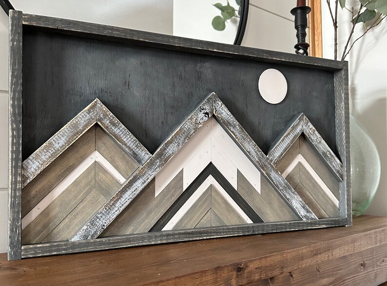 May include: A framed wooden wall art piece featuring a geometric mountain range design in shades of gray, white, and brown. The mountains are layered and textured, creating a three-dimensional effect. A white circle is centered at the top of the piece, representing the moon.