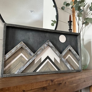 May include: A framed wood wall art piece depicting a mountain range with a white moon in the background. The mountains are made of wood strips in shades of gray, white, and brown. The frame is black.