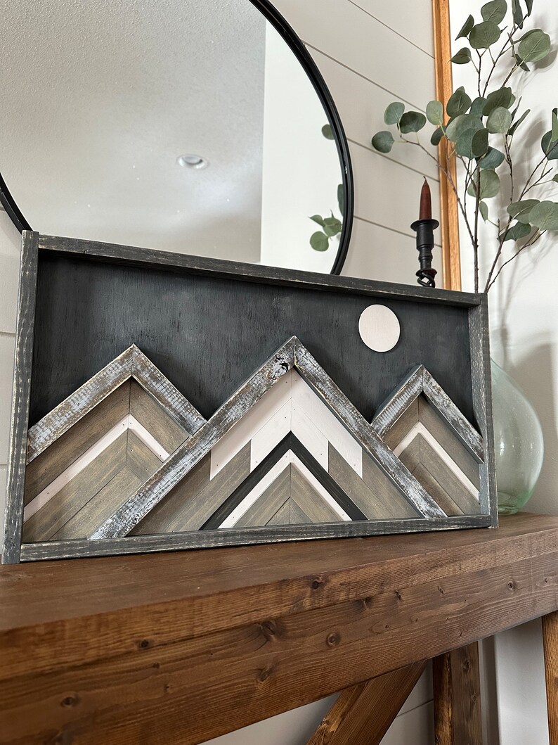 May include: A framed wood wall art piece depicting a mountain range with a white moon in the background. The mountains are made of wood strips in shades of gray, white, and brown. The frame is black.