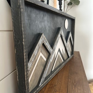 May include: A black framed wooden wall art piece with a geometric mountain design in shades of gray, white, and brown. The mountains are layered and have a distressed look. A white circle is in the upper right corner of the piece.