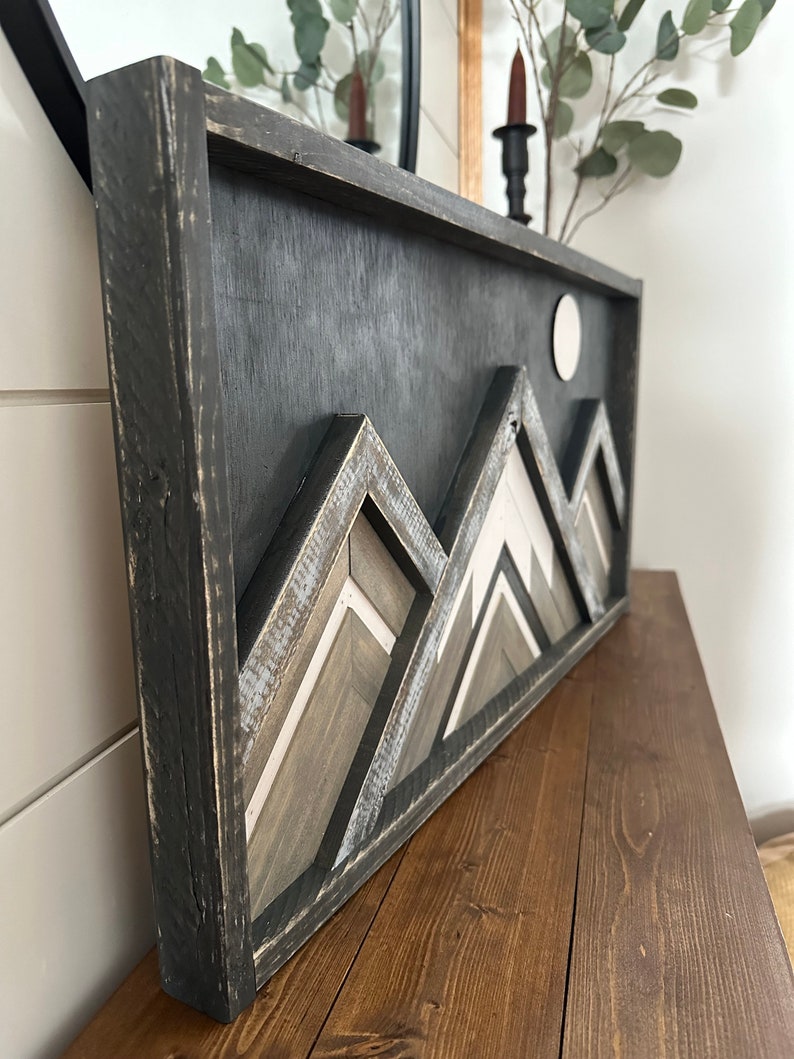 May include: A black framed wooden wall art piece with a geometric mountain design in shades of gray, white, and brown. The mountains are layered and have a distressed look. A white circle is in the upper right corner of the piece.