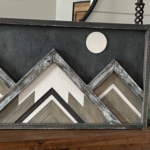 May include: A framed wood wall art piece depicting a mountain range with a white moon in the background. The mountains are made of various shades of brown and gray wood, and the frame is black.