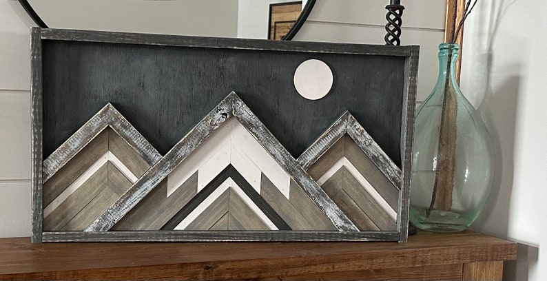 May include: A framed wood wall art piece depicting a mountain range with a white moon in the background. The mountains are made of various shades of brown and gray wood, and the frame is black.