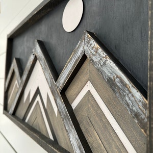 May include: A framed wood wall art piece with a black background and a white circle in the upper center. The piece features a layered design of geometric shapes in shades of gray and white, resembling mountains.