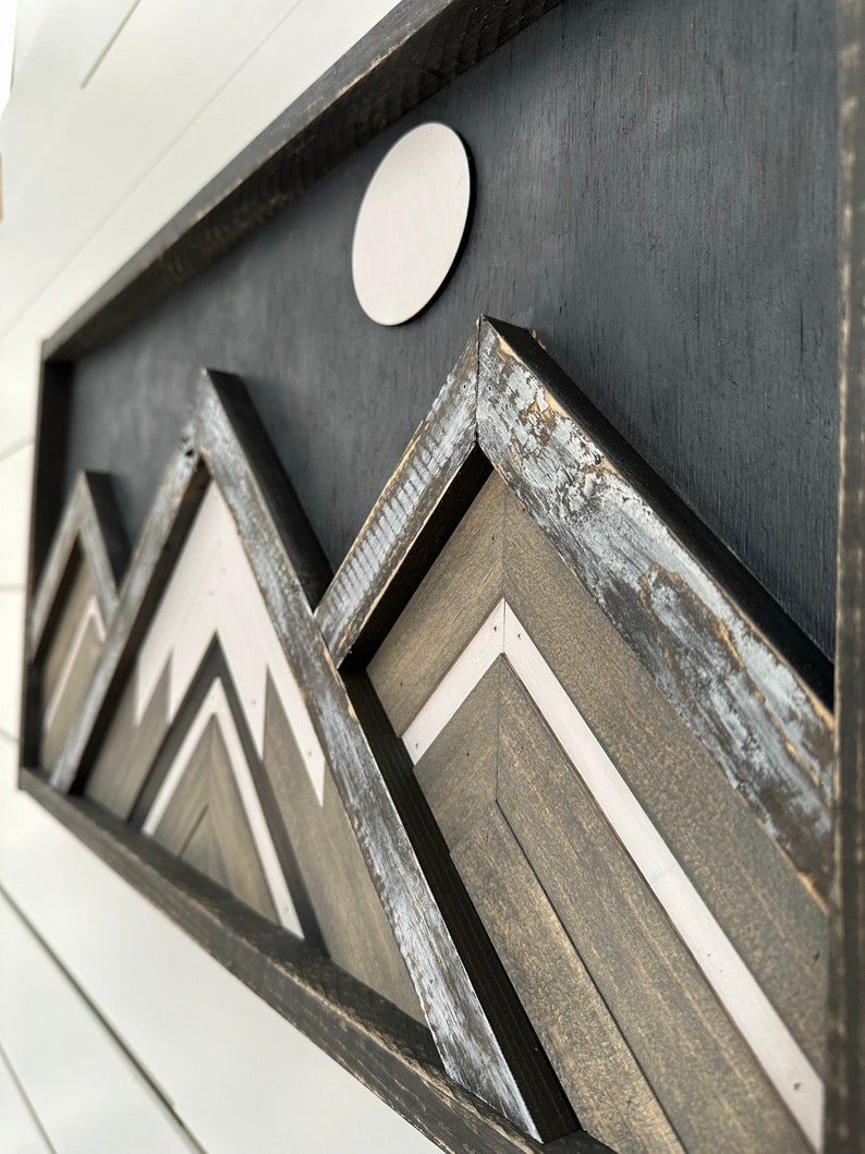 May include: A framed wood wall art piece with a black background and a white circle in the upper center. The piece features a layered design of geometric shapes in shades of gray and white, resembling mountains.