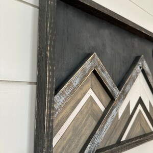 May include: A framed wooden wall art piece with a geometric mountain design. The mountains are made of wood planks in shades of brown, white, and gray. The frame is black and distressed.