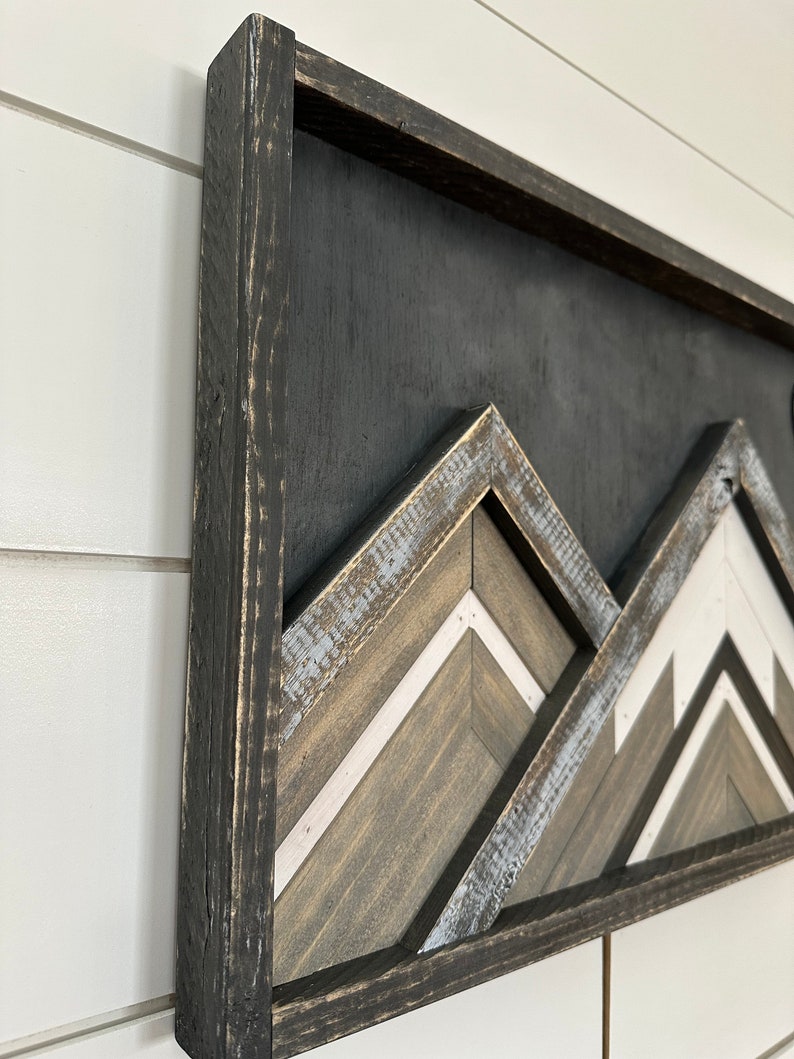 May include: A framed wooden wall art piece with a geometric mountain design. The mountains are made of wood planks in shades of brown, white, and gray. The frame is black and distressed.