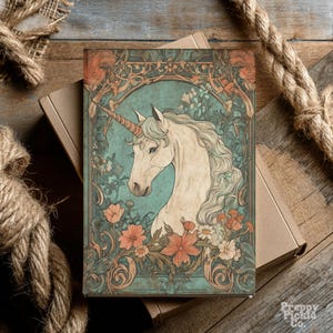 May include: A journal featuring a vintage illustration of a white unicorn with a golden horn, surrounded by flowers and foliage. The design has a teal background and is framed with ornate details. The journal is resting on a cardboard box.
