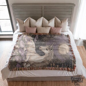 Op de afbeelding: Een geweven sprei met een gedetailleerde illustratie van een eenhoorn in een maanverlicht bos. De sprei heeft een gefranjerde rand en is voornamelijk in tinten grijs, paars en wit. Het Preppy Pickle Co. logo is rechtsonder zichtbaar.