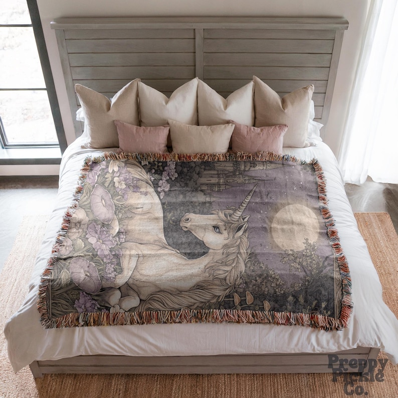 Op de afbeelding: Een geweven sprei met een gedetailleerde illustratie van een eenhoorn in een maanverlicht bos. De sprei heeft een gefranjerde rand en is voornamelijk in tinten grijs, paars en wit. Het Preppy Pickle Co. logo is rechtsonder zichtbaar.