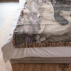 Op de afbeelding: Een geweven sprei met een gedetailleerde illustratie van een wit paard met paarse bloemen en bladeren. De deken heeft een gefranjerde rand met een mix van kleuren. Het paard ligt en het ontwerp heeft een vintage esthetiek.